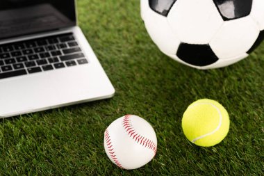 Futbol, beyzbol ve tenis topları yeşil çimlerin üzerinde dizüstü bilgisayarın yanında, spor bahisleri konsepti