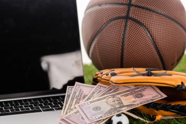 Beyazlara izole edilmiş futbol topu, dizüstü bilgisayar ve basketbol topunun yanındaki beyzbol eldivenindeki dolar banknotlarının görüntüsünü kapat.