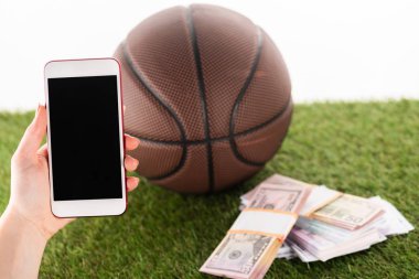 Akıllı telefonlu kadın eli, para paketlerinin yanında ve beyaz çimenlerin üzerinde izole edilmiş basketbol topu, spor bahisleri konsepti