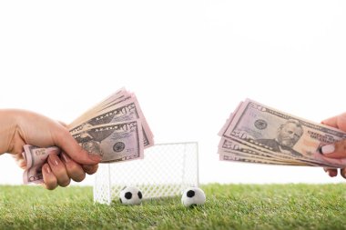 cropped view of female hands with dollar banknotes near toy soccer balls and gates on green grass isolated on white, sports betting concept