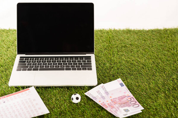 toy soccer ball, euro banknotes, betting lists near laptop with blank screen on green grass isolated on white, sports betting concept