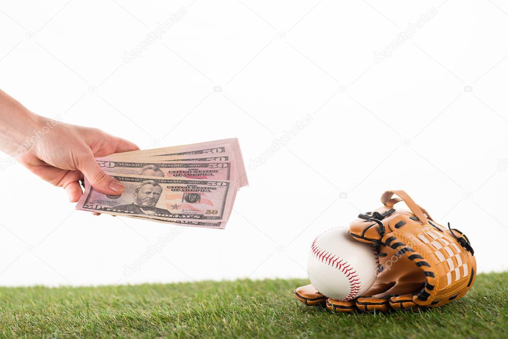 cropped view of female hand with dollar banknotes near baseball glove and ball on green grass isolated on white, sports betting concept