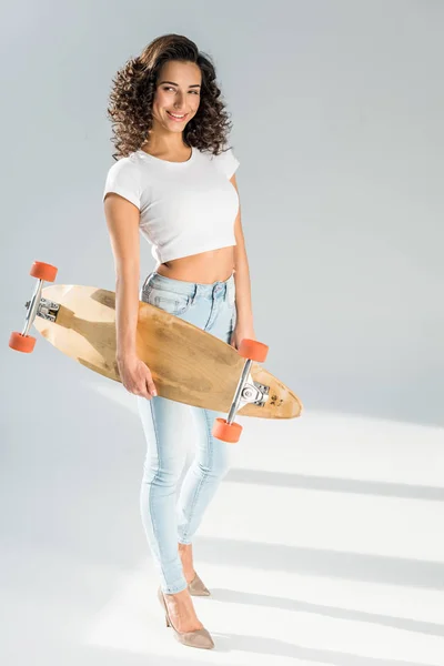 Femme gaie avec les cheveux bouclés tenant skateboard sur fond gris — Photo de stock