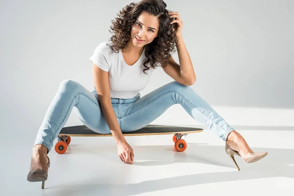 Sexy donna con i capelli ricci seduta su skateboard in tacchi alti su sfondo grigio — Foto stock