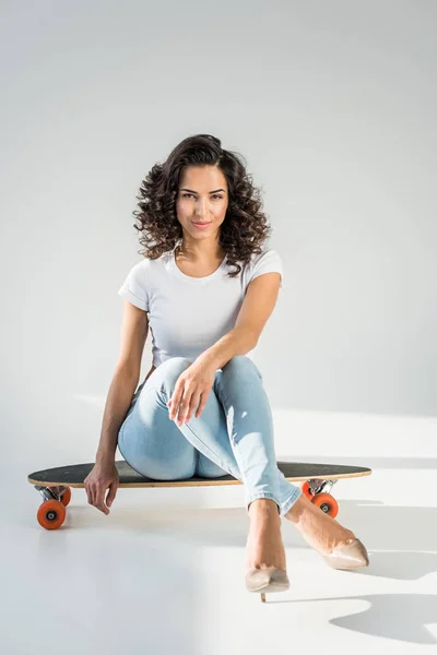 Приваблива жінка з кучерявим волоссям, що сидить на скейтборді з схрещеними ногами на сірому фоні — стокове фото
