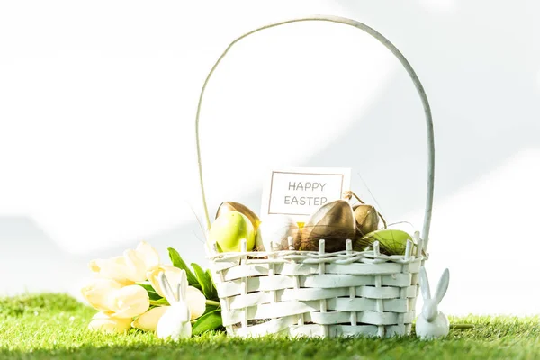 Panier de paille avec oeufs de Pâques colorés, carte de Pâques heureuse, tulipes et lapin décoratif sur blanc — Photo de stock