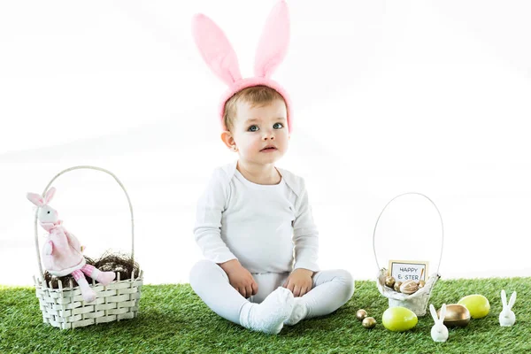 Cute baby in bunny ears headband sitting near straw baskets with Easter colorful eggs and decorative rabbits isolated on white — Stock Photo