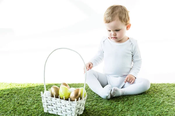 Lindo niño sentado cerca de la cesta de paja con coloridos huevos de Pascua aislados en blanco - foto de stock