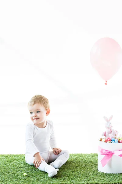 Smiling baby sitting near box with Estern eggs, decorative rabbit and air balloon isolated on white — Stock Photo