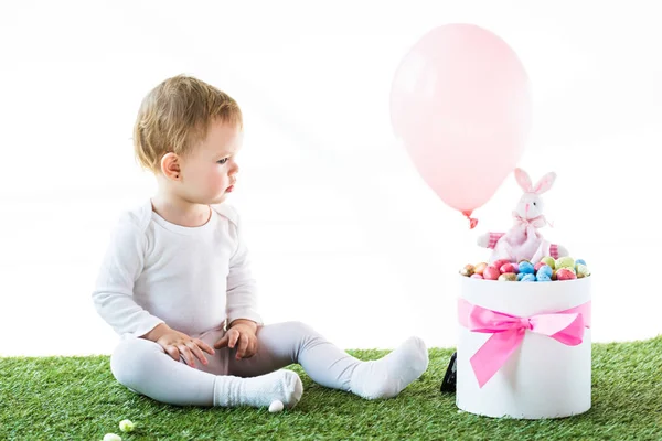 Lindo bebé sentado sobre hierba verde y mirando a la caja con huevos de Estern, conejo decorativo y globo de aire aislado en blanco - foto de stock