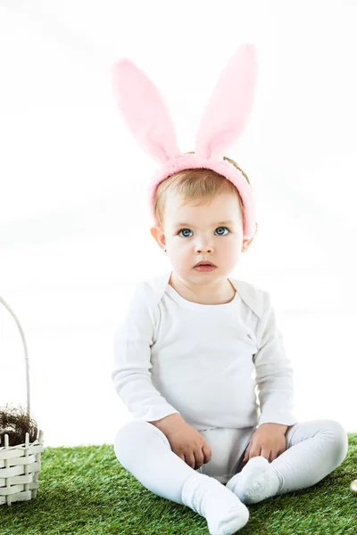 Lindo niño de ensueño en orejas de conejo diadema sentado en hierba verde cerca de la cesta de paja aislado en blanco - foto de stock