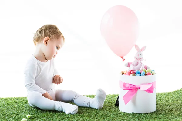 Cute baby sitting near box with Estern eggs, decorative rabbit and air balloon isolated on white — Stock Photo