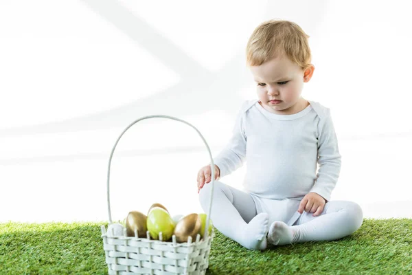 Cute baby sitting on green grass near straw basket with traditional Ester eggs isolated on white — Stock Photo