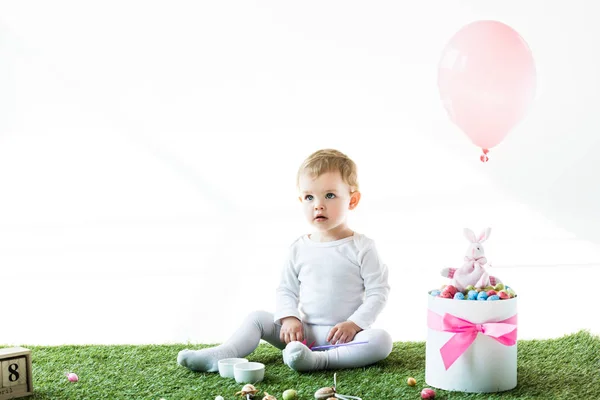 Adorable niño sentado en hierba verde cerca de la caja con huevos de codorniz de colores, conejo decorativo y globo de aire aislado en blanco - foto de stock