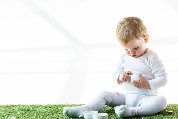 Cute baby holding chicken egg while sitting on green grass near white bowls isolated on white — Stock Photo