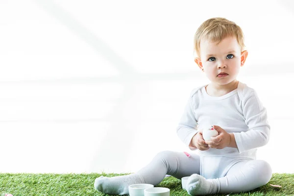 Entzückendes Baby sitzt auf grünem Gras in der Nähe weißer Schalen und hält Hühnerei isoliert auf weißer Erde — Stockfoto