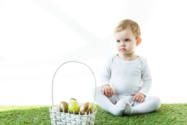Cute baby sitting on green grass near straw basket with colorful Eastern eggs isolated on white — Stock Photo