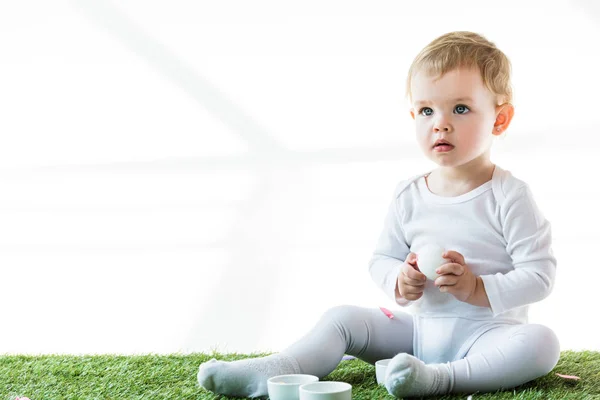 Adorable bébé tenant oeuf de poulet tout en étant assis sur l'herbe verte isolé sur blanc — Photo de stock