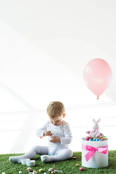 Mignon bébé assis près de la boîte avec des œufs de caille colorés, lapin décoratif et ballon d'air isolé sur blanc — Photo de stock
