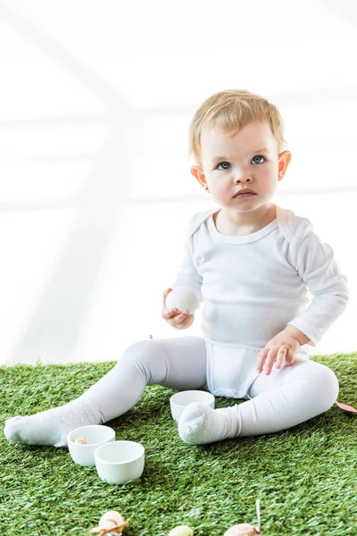 Entzückendes Kind, das Hühnerei hält, während es auf grünem Gras isoliert auf weißem Grund sitzt — Stockfoto