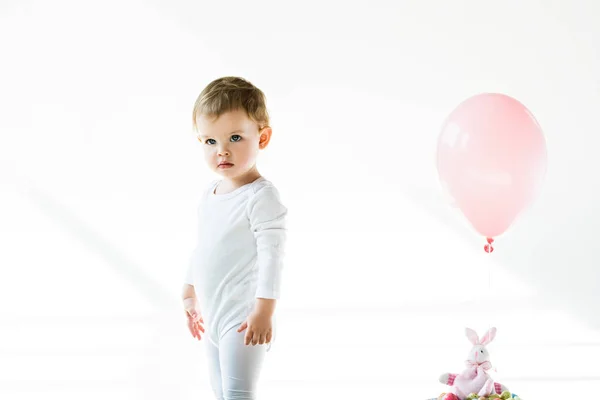 Adorabile bambino in piedi vicino coniglietto giocattolo con palloncino rosa isolato su bianco — Foto stock