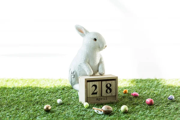 Conejo decorativo, calendario de madera con fecha 28 de abril, y coloridos huevos de Pascua sobre hierba verde aislados en blanco - foto de stock