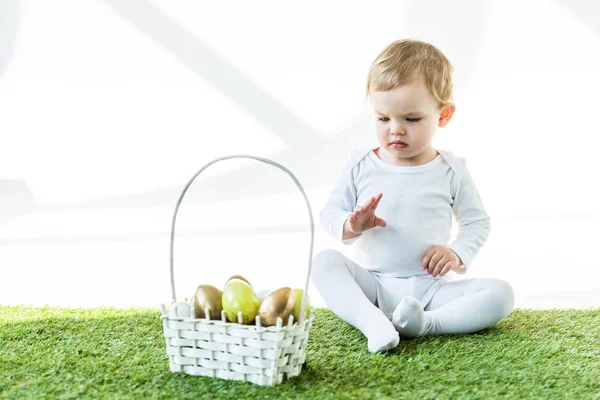 Adorable child sitting on green grass near straw basket with traditional Easter eggs isolated on white — Stock Photo