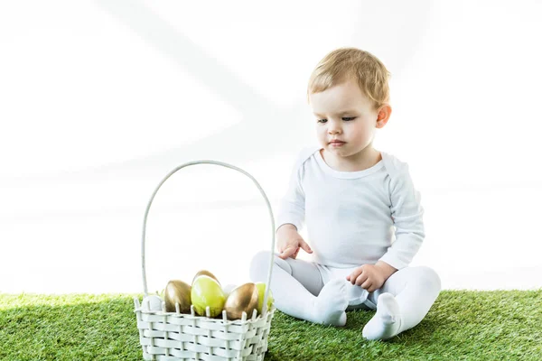 Criança bonito sentado perto da cesta de palha com ovos de Páscoa coloridos isolados em branco — Fotografia de Stock