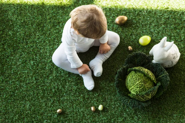 Vista aerea del bambino carino seduto sull'erba verde vicino alle uova di Pasqua, coniglio decorativo e verza — Foto stock