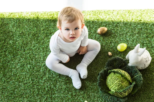 Vista aérea del adorable niño mirando a la cámara mientras está sentado cerca de los huevos de Pascua, conejo decorativo y col de col rizada — Stock Photo