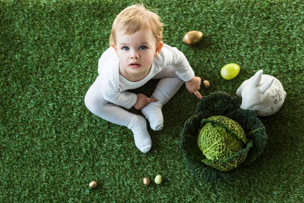 Overhead view of cute kid sitting near Easter eggs, decorative rabbit and savoy cabbage, and looking at camera — Stock Photo