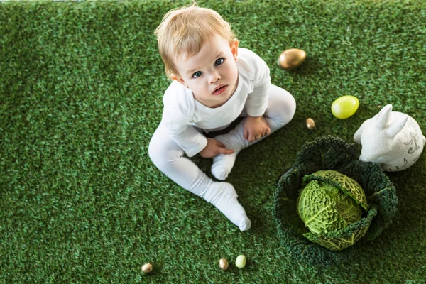 Overhead view of adorable child sitting near Easter eggs, decorative rabbit and savoy cabbage, and looking at camera — Stock Photo