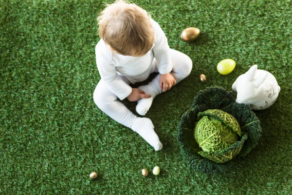 Vista superior del bebé rubio sentado en la hierba verde cerca de los huevos de Pascua, conejo decorativo y col de savoy - foto de stock