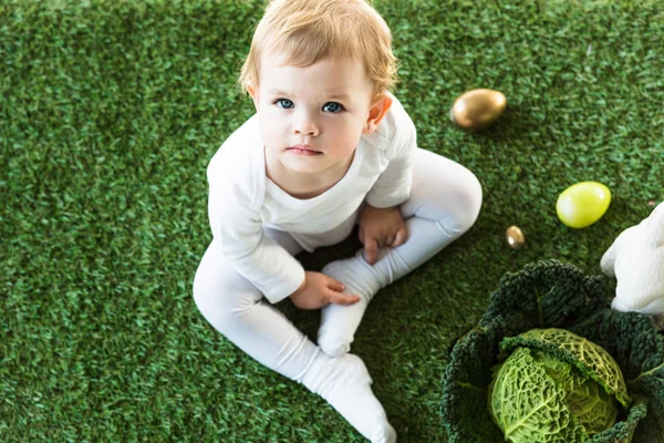 Top view of adorable child looking at camera while sitting near Easter eggs, decorative rabbit and savoy cabbage — Stock Photo