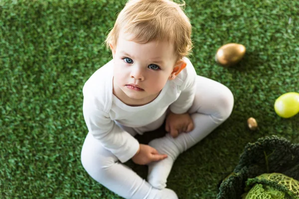 Мечтательный ребенок сидит на зеленой траве возле пасхальных яиц и савойской капусты, и смотрит в камеру — стоковое фото