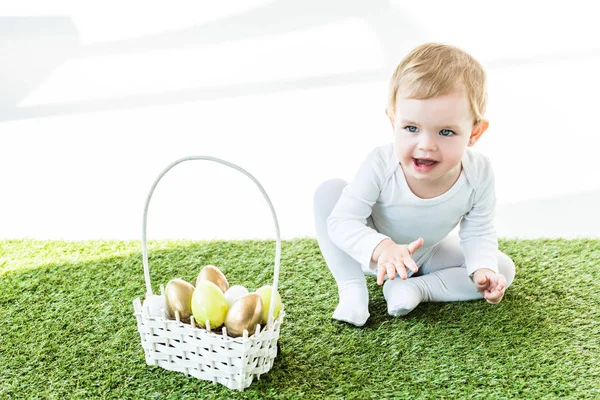 Enfant heureux assis près du panier de paille avec des œufs de Pâques colorés isolés sur blanc — Photo de stock