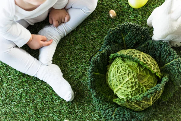 Vue recadrée de l'enfant assis sur de l'herbe verte près des œufs de Pâques, du chou de Savoie et du lapin décoratif — Photo de stock