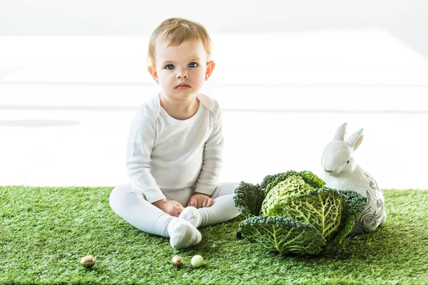 Mignon bébé assis près du chou de Savoie, lapin décoratif et oeufs de caille colorés sur blanc — Photo de stock