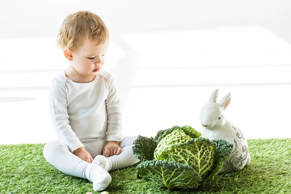 Adorabile bambino seduto su erba verde vicino verza e coniglio decorativo su bianco — Foto stock