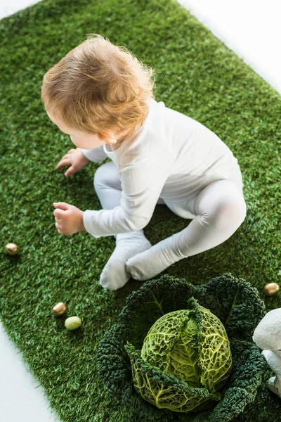 Top view of cute baby sitting on green grass near golden quail eggs and savoy cabbage — Stock Photo