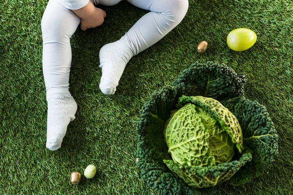Vista cortada do bebê sentado na grama verde perto de ovos de Páscoa e couve lombarda — Fotografia de Stock