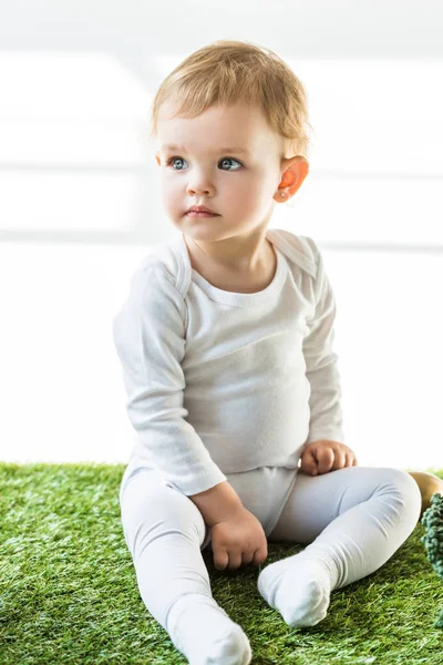 Adorable bébé avec des cheveux blonds assis sur l'herbe verte et regardant loin sur blanc — Photo de stock