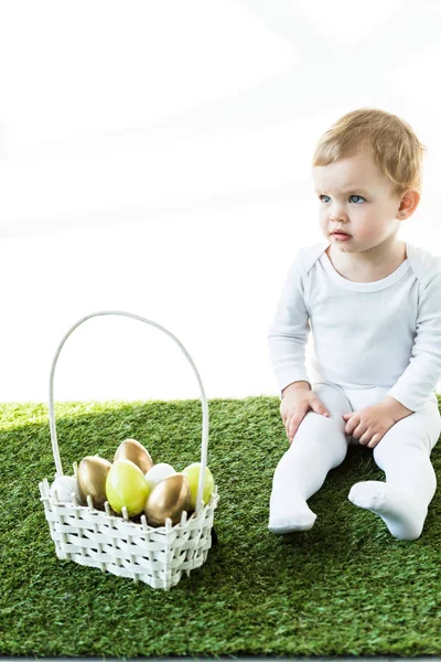 Niedliches Kind sitzt in der Nähe von Strohkorb mit bunten Ostereiern isoliert auf weiß — Stockfoto