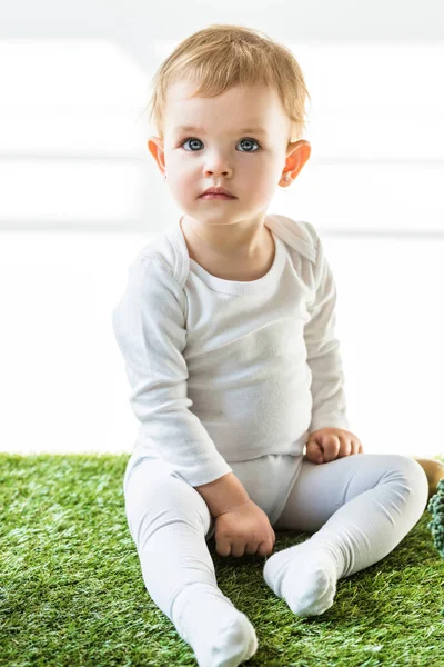 Mignon enfant avec des cheveux blonds assis sur l'herbe verte et regardant loin sur blanc — Photo de stock