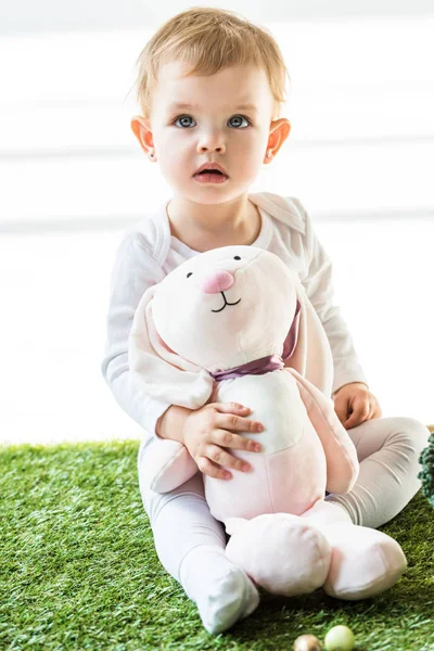 Bonito sonhador sentado no verde grama e segurando brinquedo coelho no branco — Fotografia de Stock