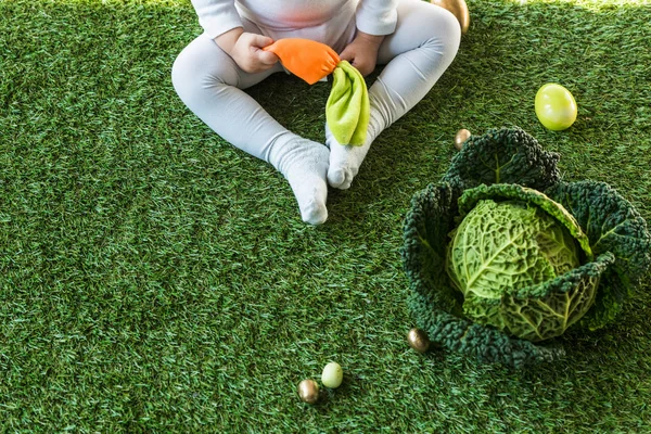 Partial view of child holding toy carrot while sitting near Easter eggs and savoy cabbage on green grass — Stock Photo