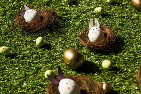 Colorful Easter eggs, and chicken eggs with funny rabbit faces and paper ears on green grass — Stock Photo