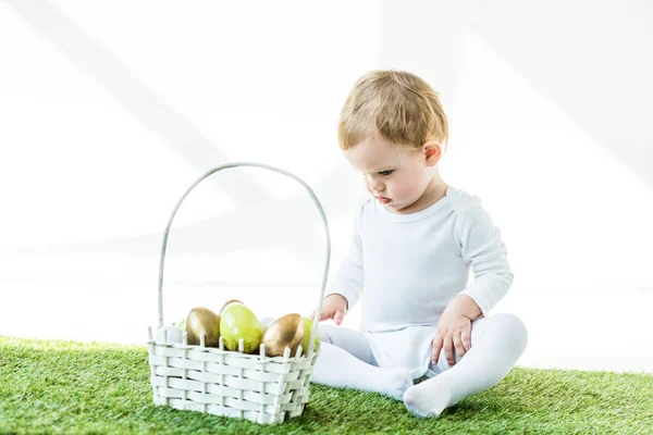 Adorabile bambino seduto vicino a cesto di paglia con uova di Pasqua lucide isolate su bianco — Foto stock