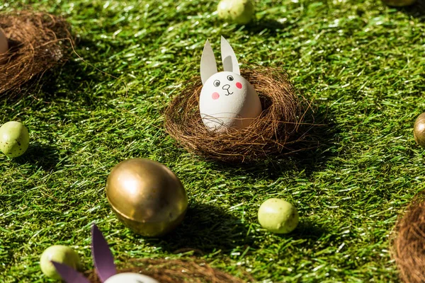 Uova di Pasqua e uova di pollo con divertente faccia di coniglio e orecchie di carta su erba verde — Foto stock