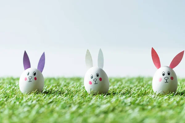 Foyer sélectif des œufs de poulet de l'Est avec des visages de lapin drôles et des oreilles en papier sur l'herbe verte — Photo de stock
